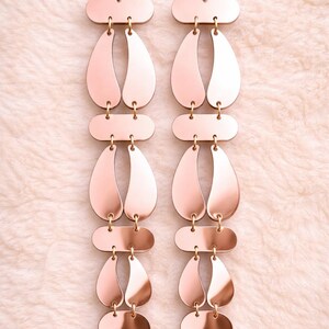 May include: A pair of long, dangling earrings with a rose gold finish. Each earring features a series of connected geometric shapes: an oval, a teardrop, and a smaller oval, all linked by small gold-colored connectors. The earrings are displayed against a soft, light pink background.