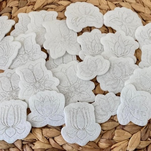May include: A collection of white, die-cut felt flower shapes with gray outlines, scattered on a woven brown surface. The floral designs include lotus and other stylized blossoms, suitable for crafting or embellishment.