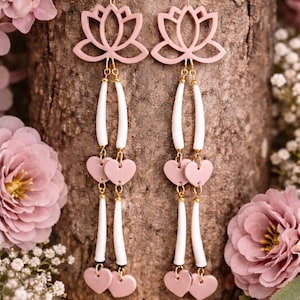 May include: Pink lotus flower earrings with white and pink heart accents. The earrings have a gold-colored hook and are set against a wooden background with pink flowers.