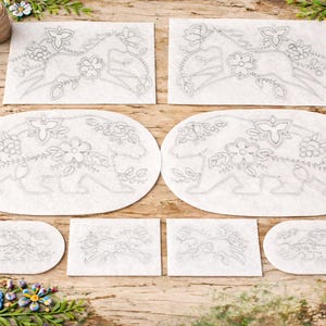 Può includere: Una collezione di modelli di ricamo con motivi floreali e orsi. I modelli sono stampati su tessuto bianco in varie forme, tra cui rettangoli e ovali. Rocchetti di filo e fiori decorativi circondano i modelli su una superficie di legno.