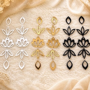 May include: Three pairs of floral-shaped earrings in silver, gold, and black. Each pair features a layered design with a teardrop-shaped bottom and a floral motif. The earrings are displayed on a cream-colored fabric.