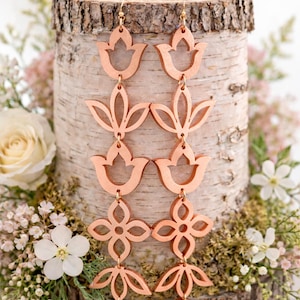 May include: Copper-colored, layered floral earrings. Each earring features multiple connected floral shapes, including tulip and four-petal designs. The earrings hang from gold-colored ear wires. The earrings are displayed against a birch log with floral accents.