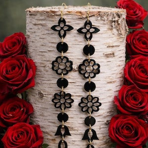Peut inclure: Boucles d'oreilles pendantes noires à motifs floraux avec crochets et connecteurs dorés. Les boucles d'oreilles présentent une série de formes connectées, dont des fleurs, des cercles et des gouttes. Les boucles d'oreilles sont présentées sur une bûche de bouleau avec des roses rouges en arrière-plan.