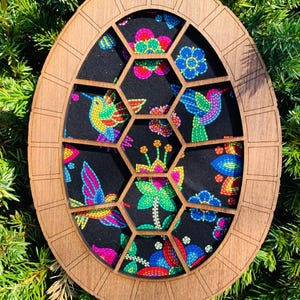 May include: Oval wooden decorative piece with a honeycomb design, showcasing a vibrant fabric with colorful hummingbird and floral patterns. The frame is a warm brown, set against a backdrop of green foliage.
