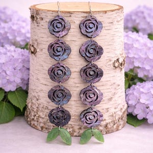 Könnte beinhalten: Ohrringe mit einem kaskadierenden Design aus dunkelvioletten rosenförmigen Anhängern mit schillernden blauen und grünen Akzenten. Jeder Ohrring hat sechs Rosenanhänger und zwei grüne Blattanhänger. Präsentiert vor einem Birkenstamm.