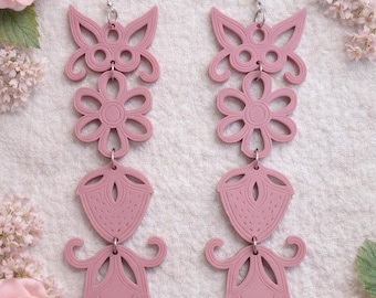 May include: Two pairs of pink, layered dangle earrings. Each earring features a butterfly shape, a flower, a teardrop shape, and a decorative bottom element. The earrings are suspended from silver-colored hooks. The background includes pink flowers.