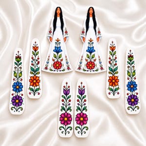 May include: Two flat figures of individuals with long black hair wearing white gowns decorated with colorful floral patterns, accompanied by six elongated white hair clips featuring vibrant hand-painted floral designs in blue, orange, purple, and green.