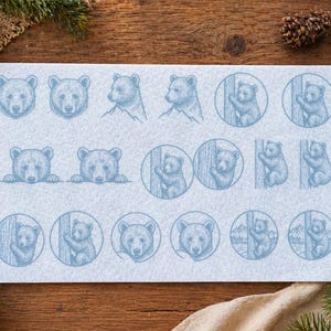 May include: A light blue felt sheet with bear illustrations. The designs include bear faces, bears with mountains, and bears climbing trees. The sheet is on a wooden surface with pinecones and greenery.