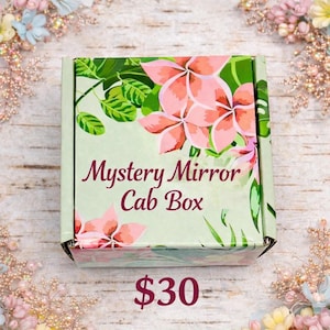 May include: A light green box with the text "Mystery Mirror Cab Box" in a cursive font. The box is decorated with pink flowers and green leaves. The box is on a light wood surface.