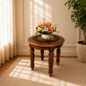 Könnte beinhalten: Ein runder Beistelltisch aus Holz mit aufwendigen Schnitzereien und einem Blumenarrangement. Der Tisch hat vier stabile Beine und eine dunkelbraune Oberfläche. Die Blumen befinden sich in einer weißen Vase und umfassen orange, rote und weiße Blüten. Der Tisch hat einen Durchmesser von etwa 61 cm.