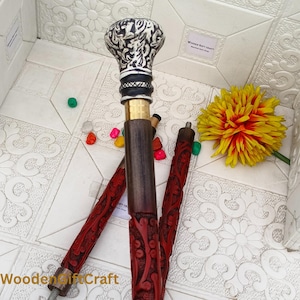 Könnte beinhalten: Dekorative Gehstock mit einem silber-schwarz gemusterten Griff. Der dunkelbraune Holzschaft ist mit Messing und einem geschnitzten roten Design akzentuiert. Der Stock ist teilweise zerlegt, die Teile werden auf einer weißen Oberfläche gezeigt.