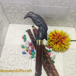 Könnte beinhalten: Ein dunkelbrauner Holzspazierstock mit einem schwarzen Rabenkopfgriff. Der Stock hat ein goldfarbenes Metallband an der Spitze und ist von anderen Holzstöcken und bunten Steinen umgeben. Der Text "WoodenGiftcraft" befindet sich unten.