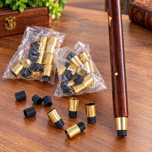 May include: A dark brown wooden walking stick with brass accents stands upright next to several small brass and black rubber cane tips. Some of the cane tips are loose on the surface, while others are contained in two clear plastic bags.