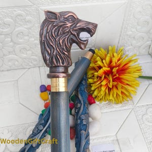 Könnte beinhalten: Dekorativer Spazierstock mit Wolfskopfgriff. Der Griff ist bronzefarben und der Schaft blau mit geschnitzten Blumenmustern. Der Stock hat einen Messingkragen und eine schwarze Spitze. Im Hintergrund befindet sich eine gelb-rote Blume.