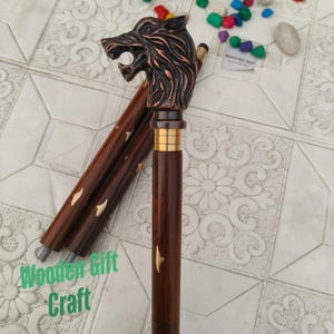 Könnte beinhalten: Ein dunkelbrauner Holzspazierstock mit einem Wolfskopfgriff. Der Wolfskopf ist bronzefarben und detailliert. Der Stock hat goldfarbene Akzente und dekorative Einlagen. Der Text "Wooden Gift Craft" ist sichtbar.