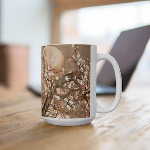 May include: White ceramic mug featuring a detailed illustration of a flowering tree with a full moon in the background. The artwork includes white blossoms and branches against a sepia-toned backdrop, creating a serene and artistic design.