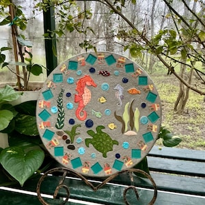 May include: A round, gray mosaic stepping stone featuring an underwater scene. The design includes a seahorse, turtle, fish, and seaweed, with turquoise and orange accents. The stone is displayed on a decorative metal stand.