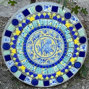 May include: A circular mosaic stepping stone with a blue and yellow floral design in the center. The stone is made of small pieces of tile and glass in various shapes and sizes. The design is surrounded by a border of blue and yellow tiles.