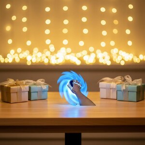 May include: A hand holding a blue and white light-up device with a swirling design. The device is emitting a bright blue light. There are gift boxes with ribbons on a wooden table. The background has a bokeh effect.
