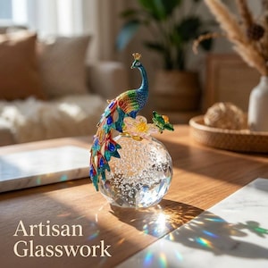 Pode incluir: Um ornamento decorativo de vidro com um pavão esmaltado colorido, pousado sobre uma esfera de cristal transparente facetada e cheia de bolhas. O pavão exibe uma plumagem vibrante em azul, verde e vermelho com detalhes dourados. Texto: Artisan Glasswork.