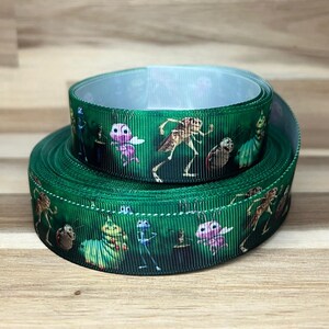 May include: A green grosgrain ribbon roll featuring a repeating pattern of various colorful insect characters from the movie A Bug's Life, including a green caterpillar, a pink butterfly, and a brown grasshopper, printed on the textured fabric surface.