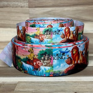 May include: Two rolls of grosgrain ribbon featuring a colorful printed pattern of characters from The Lion King, including Simba, Timon, and Pumbaa, set against a landscape background with the text Lion King printed in black on the ribbon surface.