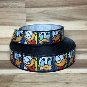 May include: A roll of black grosgrain ribbon featuring a repeating comic book style print of Donald Duck faces in blue, yellow, and white squares. The ribbon has a textured surface and is wound into a circular spool, displaying multiple character expressions.