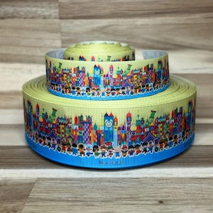 May include: Two stacked rolls of light yellow grosgrain ribbon featuring a colorful, repeating pattern of stylized buildings, clock towers, and diverse figures in bright shades of red, blue, green, and purple, finished with a solid blue bottom edge.