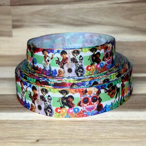 May include: A roll of grosgrain ribbon featuring a repeating pattern of characters and guitars from the movie Coco, including colorful sugar skulls and the word Coco printed in bright letters against a light green and white striped background.