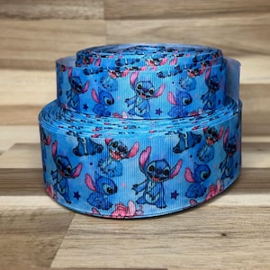 May include: A roll of light blue grosgrain ribbon featuring a repeating pattern of the blue alien character Stitch with pink ears, interspersed with small dark blue stars and pink circular accents across the textured surface of the fabric.