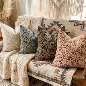 May include: A selection of patterned throw cushions in beige, brown, and black animal prints. The cushions are arranged on a wooden bench, complemented by a cream knitted blanket and a patterned rug, creating a cosy, bohemian-inspired living space.