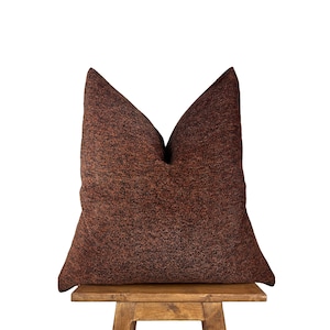 May include: A brown and black patterned throw pillow with a square shape. The pillow is sitting on a wooden stool.
