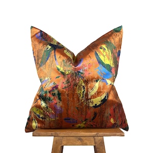May include: An orange decorative cushion with a colourful abstract design. The cushion features a mix of colours including yellow, blue, green, and pink, creating a vibrant and artistic look. The cushion is placed on a wooden stool.