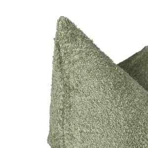 May include: Close-up of a sage green decorative cushion with a textured, looped fabric. The cushion has a soft, plush appearance and a pointed corner. The neutral colour and texture suggest a cosy home decor item.