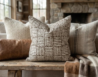 May include: A square throw pillow featuring a textured, woven checkerboard pattern in cream and dark brown. The pillow is displayed alongside a brown leather lumbar pillow, a cream knit pillow, and a striped linen pillow on a wooden bench.