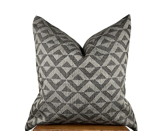May include: A decorative throw pillow with a geometric pattern in shades of gray and black. The pillow is square-shaped and rests on a small wooden stool. The pattern consists of interlocking shapes, creating a textured appearance.