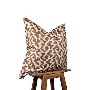 May include: A cushion with a geometric pattern in brown and cream. The cushion is placed on a small wooden stool, highlighting its design and texture. The pattern is a repeating, abstract design. The cushion is a decorative item.