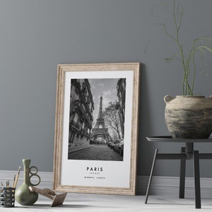 May include: Black and white photograph of the Eiffel Tower in Paris, France. The photo is framed in a light wood frame and is hanging on a gray wall.