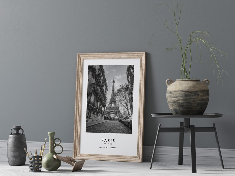 May include: Black and white photograph of the Eiffel Tower in Paris, France. The photo is framed in a light wood frame and is hanging on a gray wall.
