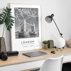 May include: A black and white framed print of the Tower Bridge in London, England, on a wooden desk. The desk also holds a black desk lamp, a laptop, a camera, a speaker, a moon-shaped lamp, and a white chair.