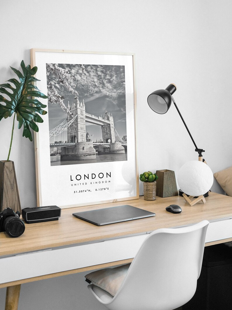 May include: A black and white framed print of the Tower Bridge in London, England, on a wooden desk. The desk also holds a black desk lamp, a laptop, a camera, a speaker, a moon-shaped lamp, and a white chair.
