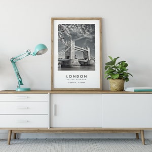 May include: A framed black and white print of the Tower Bridge in London, with the city name and coordinates below. The print is displayed on a white and wood-toned cabinet, alongside a teal desk lamp and potted plants.