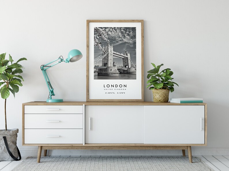 May include: A framed black and white print of the Tower Bridge in London, with the city name and coordinates below. The print is displayed on a white and wood-toned cabinet, alongside a teal desk lamp and potted plants.