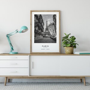 May include: A black and white photograph of the Eiffel Tower in Paris, France. The photo is framed in a light wood frame and is displayed on a white and wood cabinet with a teal blue lamp and a potted plant.