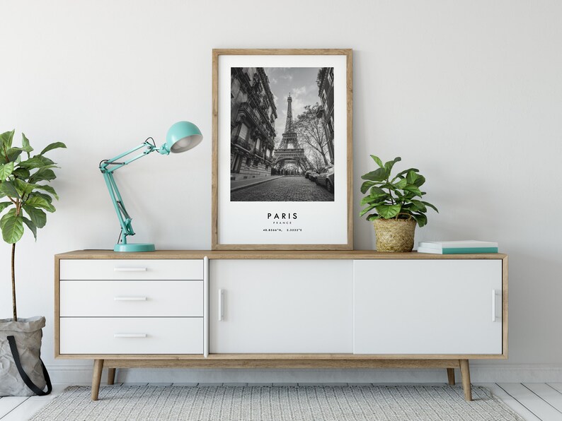 May include: A black and white photograph of the Eiffel Tower in Paris, France. The photo is framed in a light wood frame and is displayed on a white and wood cabinet with a teal blue lamp and a potted plant.