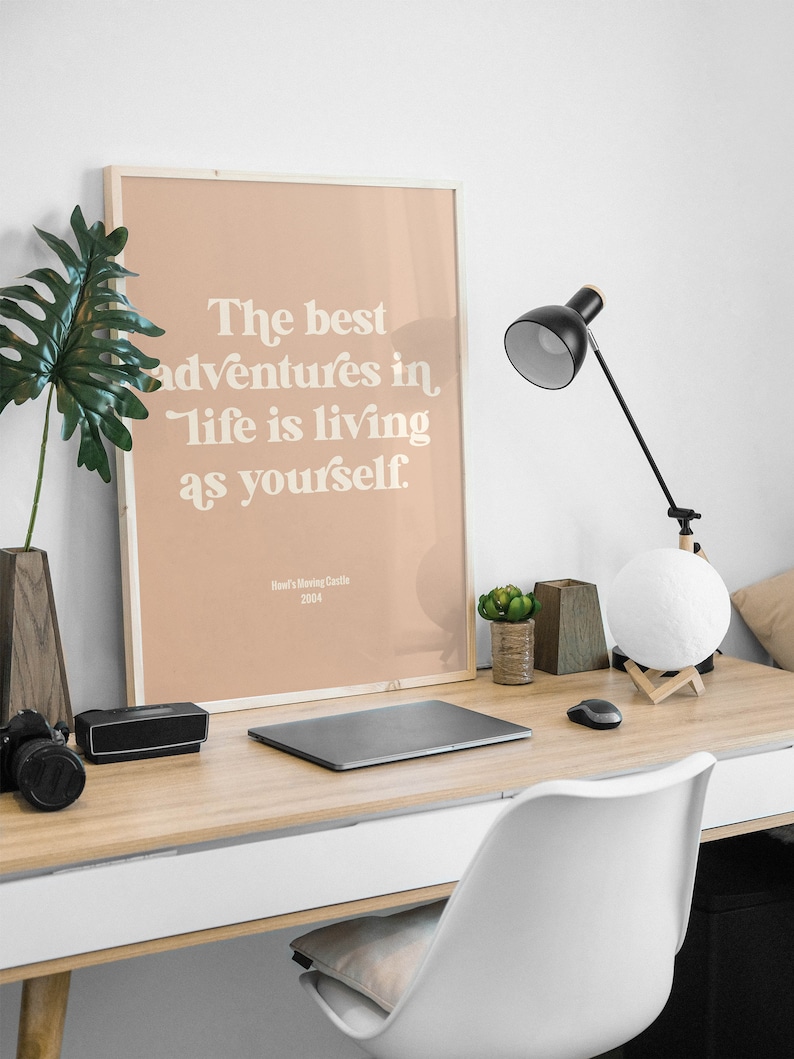 Może przedstawiać: Wydruk w ramie z brzoskwiniowym tłem i białym tekstem, kt&oacute;ry brzmi "The best adventures in life is living as yourself." Tekst jest w retro stylu czcionki. Na dole wydruku znajduje się tekst "Howl's Moving Castle 2004".