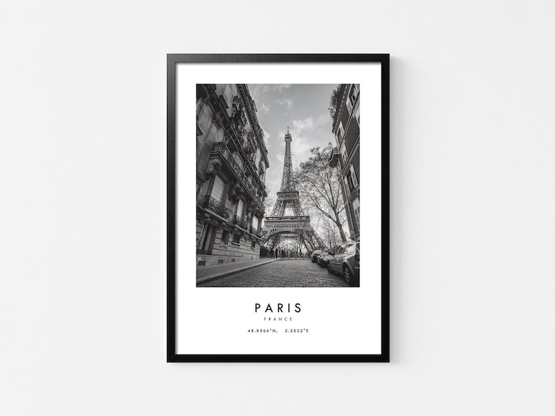 May include: Black and white photograph of the Eiffel Tower in Paris, France. The tower is seen from a street level perspective, with buildings on either side of the street. The text "PARIS, FRANCE, 48.8584&deg; N, 2.2945&deg; E" is at the bottom of the image.