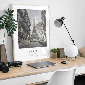May include: Black and white photograph of the Eiffel Tower in Paris, France. The photo is framed in a light wood frame and is hanging on a white wall. A desk with a laptop, a lamp, and a plant is in the foreground.