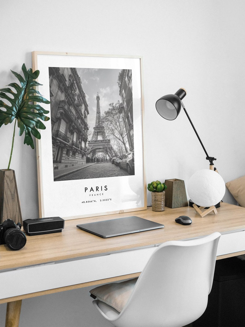 May include: Black and white photograph of the Eiffel Tower in Paris, France. The photo is framed in a light wood frame and is hanging on a white wall. A desk with a laptop, a lamp, and a plant is in the foreground.
