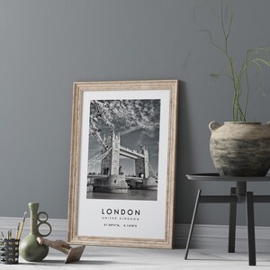 May include: A framed black and white print of the Tower Bridge in London, United Kingdom, with the city's name and coordinates. The print has a light wood frame and is leaning against a gray wall. A decorative vase and other objects are in the foreground.